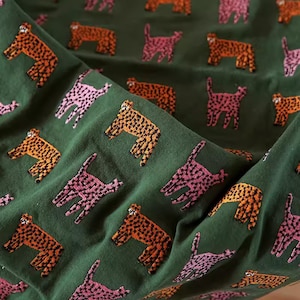 Könnte beinhalten: Grüner Stoff mit einem sich wiederholenden Muster aus pinkfarbenen und orangen, stilisierten Tierfiguren. Die Tiere sind alle in die gleiche Richtung ausgerichtet und haben ein gepunktetes Muster.