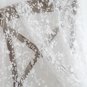 May include: White sheer fabric with a delicate floral embroidery pattern. The fabric is draped over a wooden chair.
