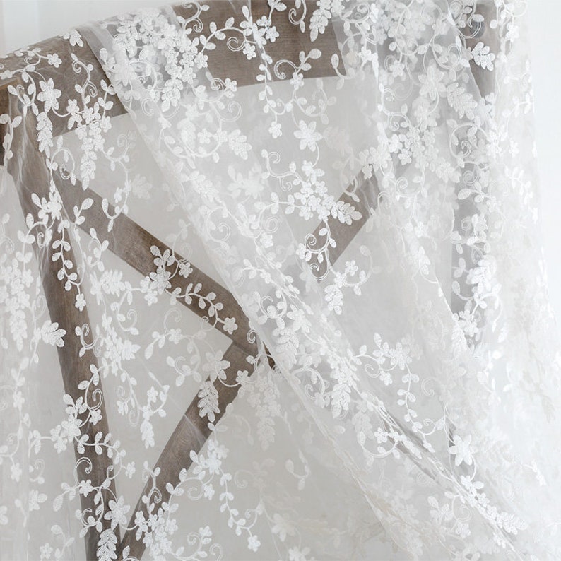 May include: White sheer fabric with a delicate floral embroidery pattern. The fabric is draped over a wooden chair.