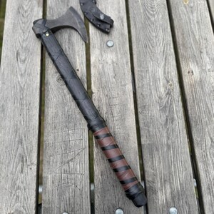 May include: A black and brown axe with a leather-wrapped handle and a metal blade. The handle is wrapped with black leather and brown leather accents. A black leather sheath is next to the axe. The axe is on a wooden surface.