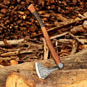 Könnte beinhalten: Axt mit Holzgriff und dekorativem, graviertem Metallkopf. Der Griff ist in der Nähe des Kopfes mit schwarzem Leder umwickelt. Die Axt liegt auf einem Baumstamm, mit einem Hintergrund aus gestapeltem Holz.
