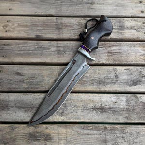 Puede incluir: Un cuchillo de hoja fija con una hoja de acero Damasco estampada y una incrustación ondulada de color cobre. El mango está hecho de madera oscura con un espaciador morado, una virola plateada y un cordón negro con una cuenta plateada.