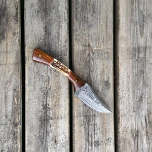 Peut inclure: Un couteau à lame fixe doté d'une lame argentée avec un motif ondulé en couches et un manche en bois brun foncé poli avec une section incrustée texturée brun clair, fixée par deux petites goupilles métalliques près de la mitre.