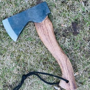 Op de afbeelding: Een kleine bijl met een donkergrijs stalen blad en een gebogen houten handvat. De handgreep is lichtbruin en heeft een zwart koord. De bijl ligt op groen gras.