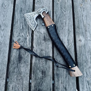 Puede incluir: Un hacha de mano con una cabeza de metal plateado que presenta intrincados patrones grabados. El mango es de madera y está parcialmente envuelto en una empuñadura de cuero negro trenzado asegurada con remaches de latón. Un cordón de cuero negro.