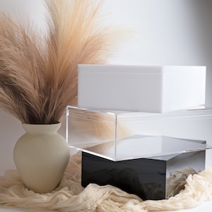 May include: Three stacked boxes, one white, one clear acrylic, and one black. The boxes are on a beige fabric surface. A vase with dried pampas grass is in the foreground.