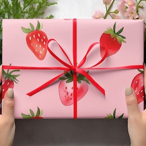 May include: A pink gift box with a red ribbon and a strawberry pattern. The box is wrapped in pink paper with red strawberries printed on it.