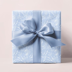 Puede incluir: Una caja de regalo azul claro con un patr&oacute;n floral blanco y una cinta de sat&eacute;n azul a juego atada en un lazo.