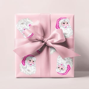 Puede incluir: Un regalo envuelto con papel de regalo rosa con ilustraciones de acuarela de caras de Pap&aacute; Noel. Una gran cinta y lazo de sat&eacute;n rosa est&aacute;n atados alrededor del regalo. Las caras de Pap&aacute; Noel tienen barbas blancas y gorros rosas.