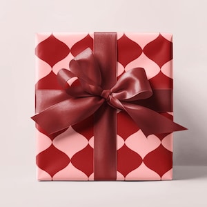 May include: A square gift box wrapped in pink paper with a repeating red teardrop pattern. A burgundy satin ribbon is tied around the box with a large bow on top. The gift is ready to be given.