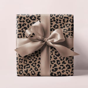 May include: A wrapped gift box featuring a leopard print design in shades of brown and black. The box is tied with a wide, satin ribbon and bow in a matching neutral color. The gift is presented on a soft pink background.