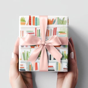 May include: A wrapped gift with a pink satin ribbon. The wrapping paper features a repeating pattern of colorful bookshelves with books and plants. The gift is held by two hands, with white sleeves and a light gray background.