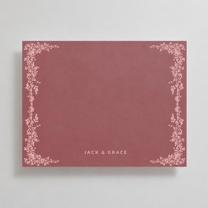 Puede incluir: Una tarjeta rectangular de color rosa polvoriento con un delicado borde de enredaderas florales de color rosa claro en los lados izquierdo y derecho. El texto "JACK & GRACE" está impreso en una fuente blanca sin remates en la parte inferior.