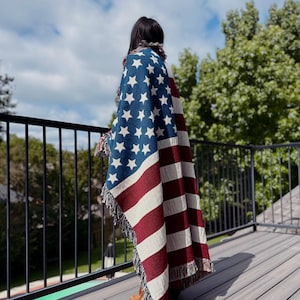 Könnte beinhalten: Eine gewebte Decke mit dem Design der amerikanischen Flagge, mit blauen Sternen und rot-weißen Streifen. Die Decke hat Fransen an den Kanten und ist über einer Person auf einer Holzterrasse drapiert, mit einem schwarzen Geländer und blauem Himmel im Hintergrund.