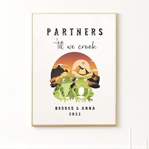 May include: A framed art print featuring two green cartoon frogs wearing cowboy hats, set against a sunset landscape. The text reads "PARTNERS 'til we croak" above the frogs, with "BROOKS & ANNA 2022" below.