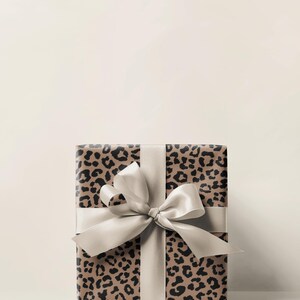 May include: A wrapped gift box featuring a leopard print design in shades of brown and black. The box is tied with a wide, light-colored satin ribbon and bow. The overall aesthetic is elegant and suitable for various occasions.