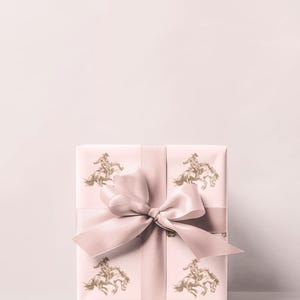 May include: A wrapped gift box with a light pink background and a matching ribbon. The wrapping paper features a repeating pattern of a gold-colored cowboy riding a horse. The bow is tied in the center.