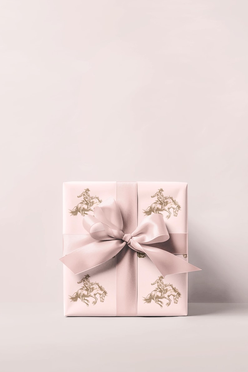 May include: A wrapped gift box with a light pink background and a matching ribbon. The wrapping paper features a repeating pattern of a gold-colored cowboy riding a horse. The bow is tied in the center.