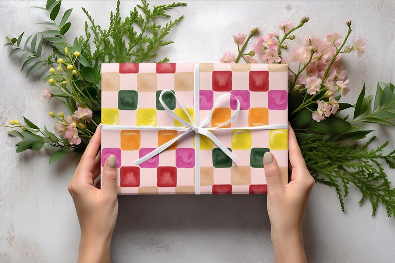K&ouml;nnte beinhalten: Eine Geschenkbox, verpackt in rosa und wei&szlig; kariertes Papier mit bunten Quadraten. Das Papier hat einen Aquarell-Effekt und die Quadrate sind mit verschiedenen Farben gef&uuml;llt, darunter Rot, Orange, Gelb, Gr&uuml;n und Blau.