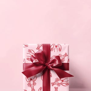 May include: A wrapped gift box with a red lobster pattern on white paper. The box is tied with a wide, burgundy ribbon and bow. The background is a solid light pink.