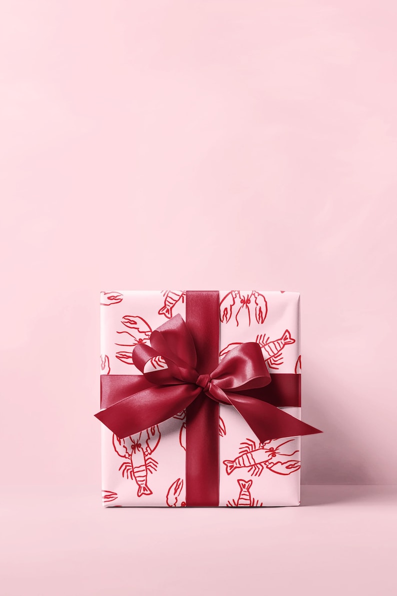 May include: A wrapped gift box with a red lobster pattern on white paper. The box is tied with a wide, burgundy ribbon and bow. The background is a solid light pink.