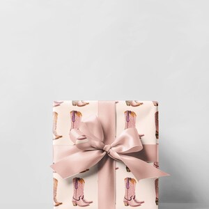 Pu&ograve; includere: Una scatola regalo avvolta in carta da regalo rosa e bianca con un fiocco rosa. La carta da regalo ha un motivo di stivali da cowboy rosa.
