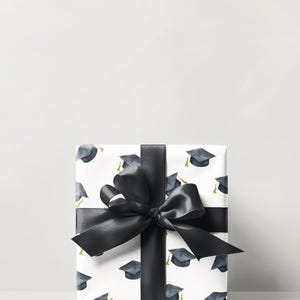 K&ouml;nnte beinhalten: Eine wei&szlig;e Geschenkbox mit schwarzem Band und Schleife. Die Box ist in Geschenkpapier mit einem Muster aus Abschlusskappen verpackt.