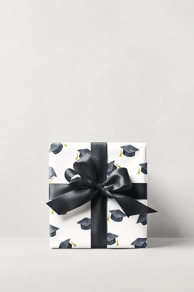 K&ouml;nnte beinhalten: Eine wei&szlig;e Geschenkbox mit schwarzem Band und Schleife. Die Box ist in Geschenkpapier mit einem Muster aus Abschlusskappen verpackt.