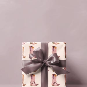 Pu&ograve; includere: Una scatola regalo avvolta in carta bianca con un motivo di stivali da cowboy rosa. La scatola &egrave; legata con un nastro di raso grigio.