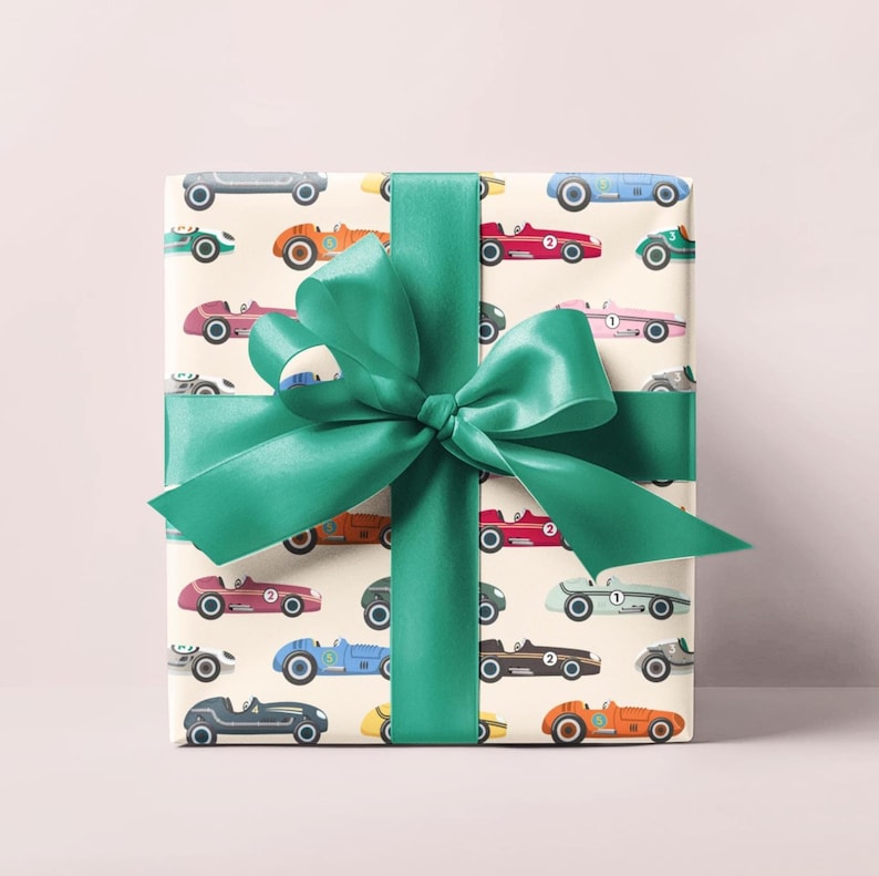May include: Gift-wrapped present featuring a cream-colored paper with a pattern of vintage race cars in various colors. The gift is tied with a large, emerald green satin ribbon and bow. The background is a soft, solid pink.