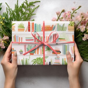 May include: A wrapped gift with a pink ribbon, featuring a white paper with a bookshelf pattern. The design includes colorful books and potted plants. Greenery and pink flowers surround the gift, creating a decorative presentation.