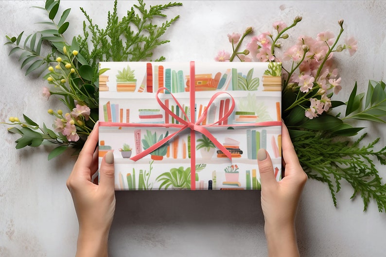 May include: A wrapped gift with a pink ribbon, featuring a white paper with a bookshelf pattern. The design includes colorful books and potted plants. Greenery and pink flowers surround the gift, creating a decorative presentation.