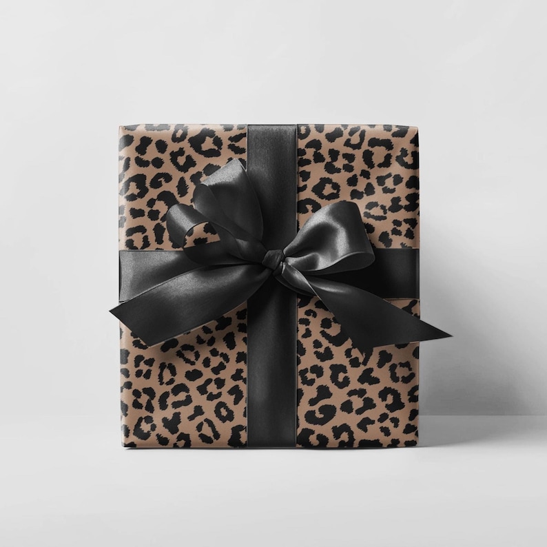 May include: A wrapped gift box featuring a tan and black leopard print pattern. The box is tied with a wide black satin ribbon and a large bow. The gift is ready to be given.