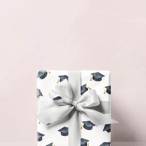 K&ouml;nnte beinhalten: Eine wei&szlig;e Geschenkbox mit einer silbernen Schleife und einem Muster aus Abschlusskappen. Die Box ist in wei&szlig;es Papier mit einem sich wiederholenden Muster aus schwarzen Abschlusskappen mit goldenen Quasten verpackt.