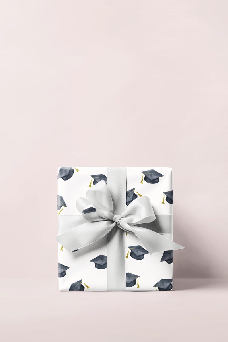 K&ouml;nnte beinhalten: Eine wei&szlig;e Geschenkbox mit einer silbernen Schleife und einem Muster aus Abschlusskappen. Die Box ist in wei&szlig;es Papier mit einem sich wiederholenden Muster aus schwarzen Abschlusskappen mit goldenen Quasten verpackt.