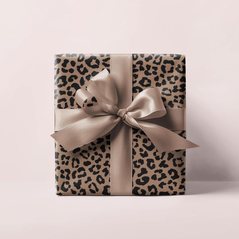 May include: A wrapped gift box featuring a leopard print design in shades of brown and black. The box is tied with a wide, satin ribbon and bow in a matching neutral color. The gift is presented on a soft pink background.