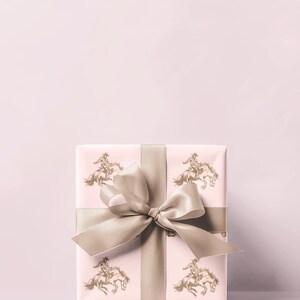 May include: A wrapped gift box with a light pink background. The wrapping paper features a repeating pattern of a rider on a horse in a gold color. A wide, light brown satin ribbon is tied around the box with a large bow.
