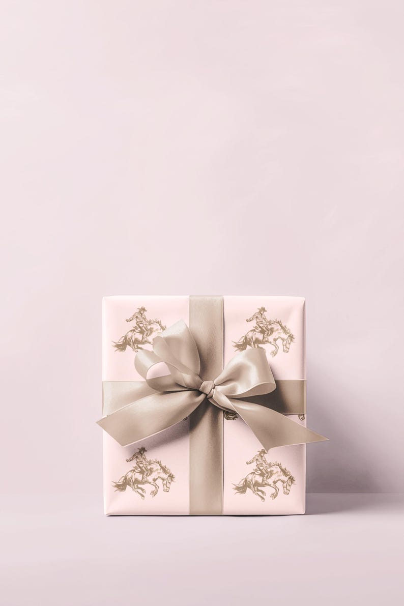 May include: A wrapped gift box with a light pink background. The wrapping paper features a repeating pattern of a rider on a horse in a gold color. A wide, light brown satin ribbon is tied around the box with a large bow.