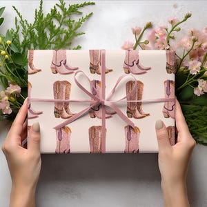Pu&ograve; includere: Un regalo avvolto in carta bianca con un nastro rosa. La carta ha un motivo ripetuto di stivali da cowboy rosa con decorazioni viola.