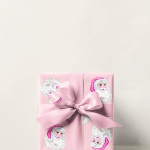 Puede incluir: Una caja de regalo rosa envuelta en papel con ilustraciones de Pap&aacute; Noel. La caja est&aacute; atada con una cinta y un lazo rosas a juego. Las ilustraciones de Pap&aacute; Noel tienen barbas blancas y gorros rosas.