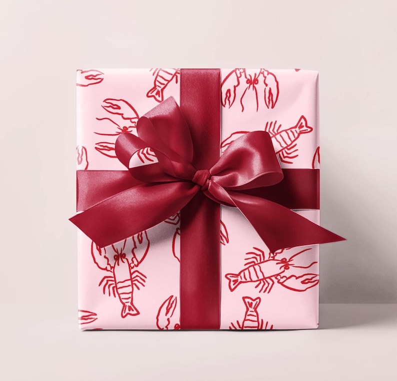 May include: A wrapped gift with light pink wrapping paper featuring a red lobster pattern. A wide, dark red satin ribbon is wrapped around the gift and tied into a large bow.