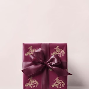 May include: A burgundy gift box wrapped in paper with a gold cowboy and horse illustration. The box is tied with a matching satin ribbon and bow. The bow is in the centre of the box.