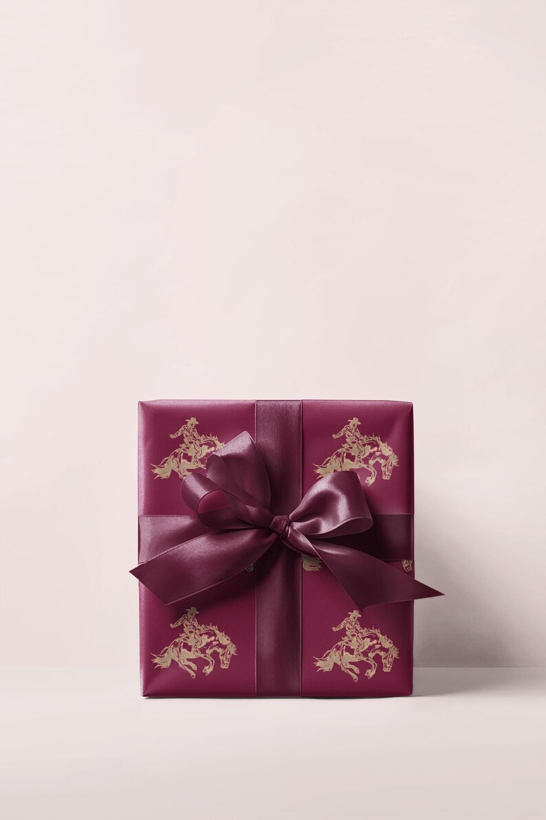 May include: A burgundy gift box wrapped in paper with a gold cowboy and horse illustration. The box is tied with a matching satin ribbon and bow. The bow is in the centre of the box.