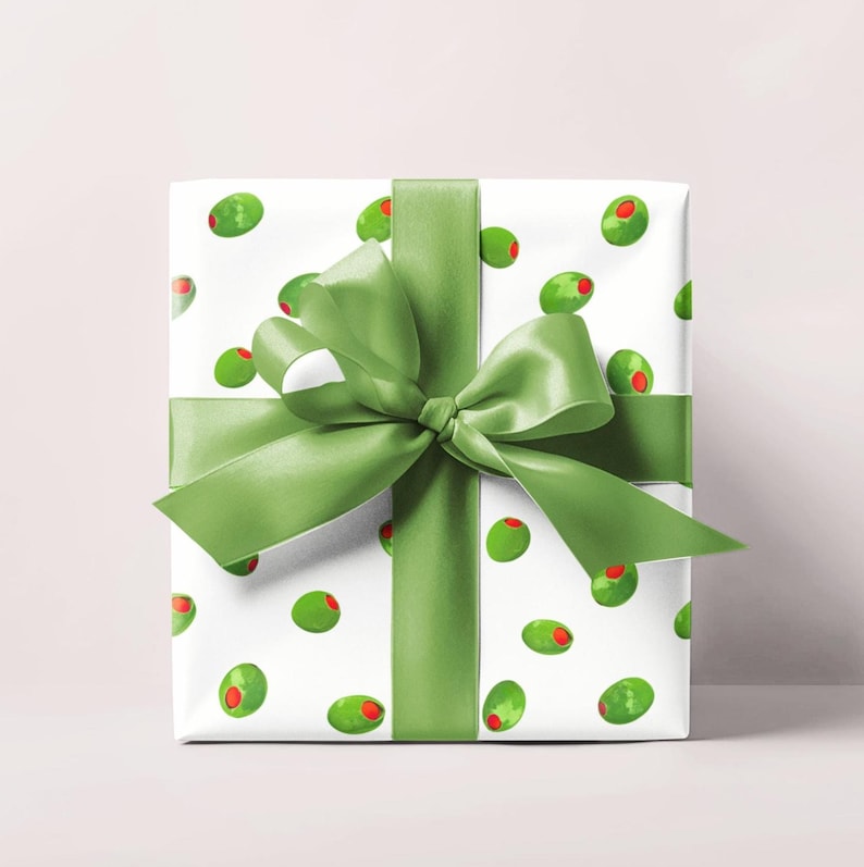May include: A wrapped gift with white paper featuring a pattern of green olives with red centers. The gift is tied with a wide, light green satin ribbon and a large bow. The background is a solid, light pink color.