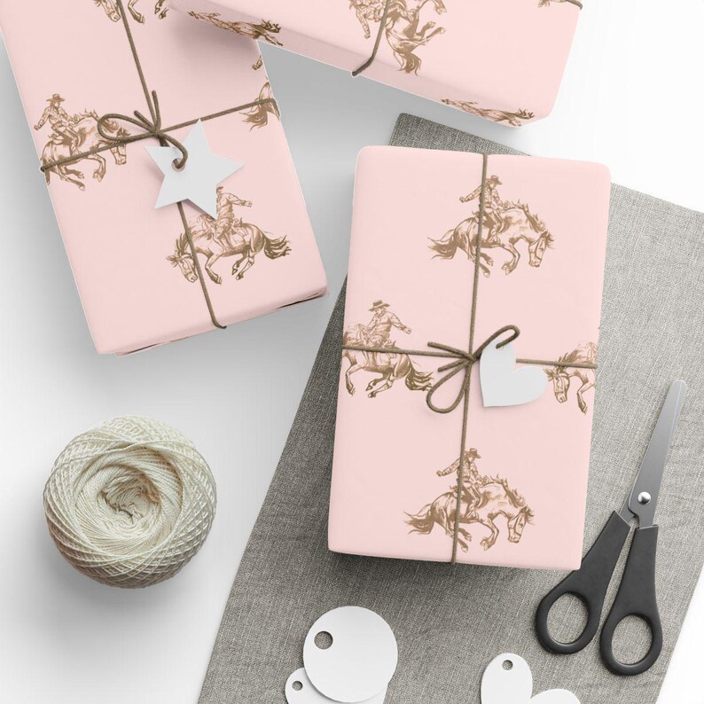 May include: Pink gift wrap featuring a repeating pattern of a cowboy riding a horse. The wrapped gifts are tied with brown twine and adorned with white star and heart-shaped tags. A ball of twine and scissors are also visible.