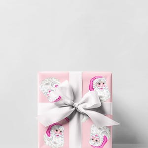 Puede incluir: Una caja de regalo envuelta con papel rosa estampado con caras de Pap&aacute; Noel. La caja est&aacute; atada con una gran cinta y lazo de sat&eacute;n blanco, ideal para Navidad o festividades.