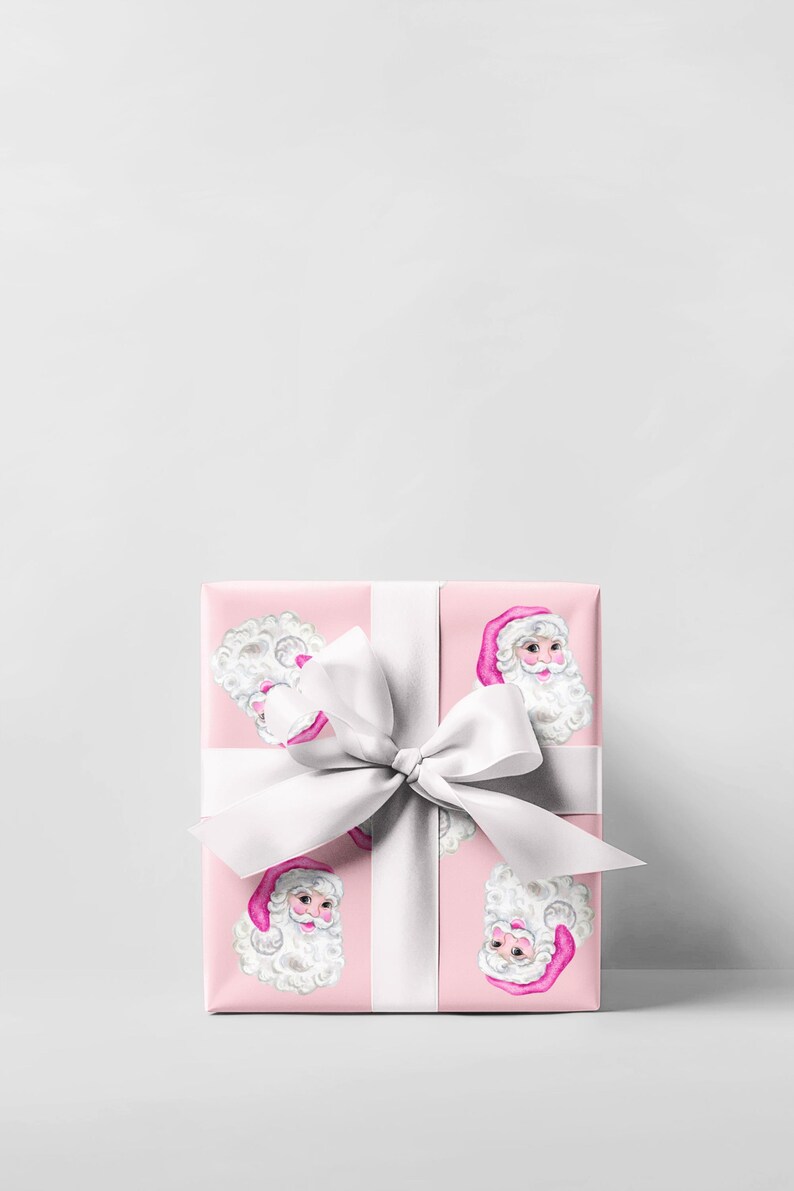 Puede incluir: Una caja de regalo envuelta con papel rosa estampado con caras de Pap&aacute; Noel. La caja est&aacute; atada con una gran cinta y lazo de sat&eacute;n blanco, ideal para Navidad o festividades.