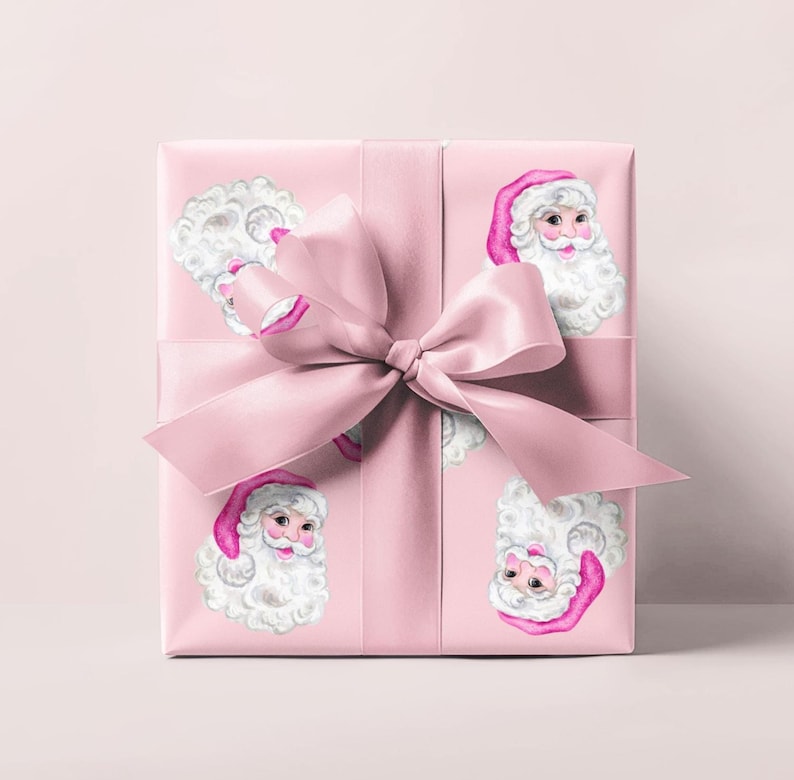 Puede incluir: Un regalo envuelto con papel de regalo rosa con ilustraciones de acuarela de caras de Pap&aacute; Noel. Una gran cinta y lazo de sat&eacute;n rosa est&aacute;n atados alrededor del regalo. Las caras de Pap&aacute; Noel tienen barbas blancas y gorros rosas.