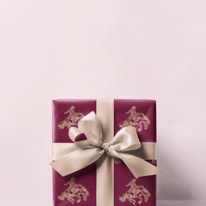 May include: A wrapped gift box with a deep red paper featuring a gold cowboy and horse pattern. The box is tied with a cream-coloured satin ribbon and bow. The background is a soft pink.