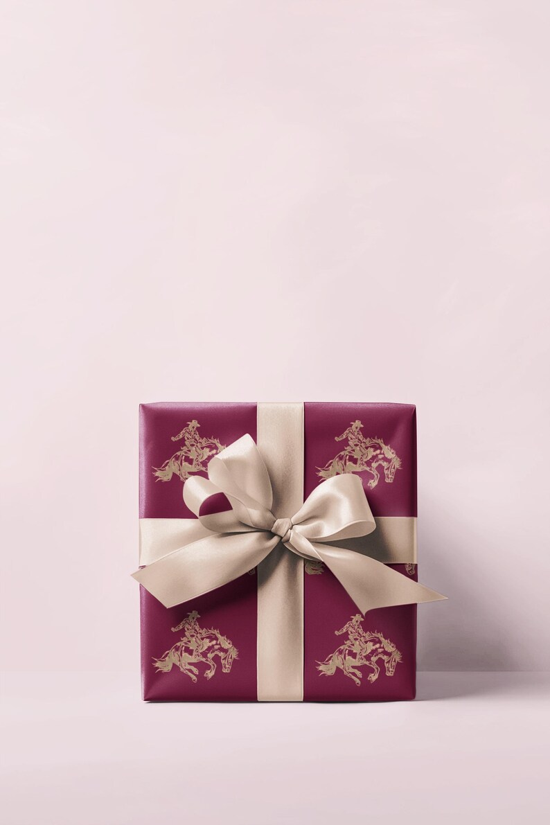 May include: A wrapped gift box with a deep red paper featuring a gold cowboy and horse pattern. The box is tied with a cream-coloured satin ribbon and bow. The background is a soft pink.